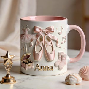 May include: White ceramic mug with a pink interior and handle, decorated with ballet-themed relief designs. Features ballet shoes, a tutu, musical notes, and the name "Anna" in gold lettering. Includes a gold star trophy and seashells.