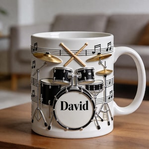 Może przedstawiać: Biały ceramiczny kubek z trójwymiarowym wzorem perkusji, z czarnymi bębnami, złotymi talerzami i pałeczkami. Imię "David" jest wyświetlane na bębnie basowym. Nuty i pięciolinie zdobią kubek.