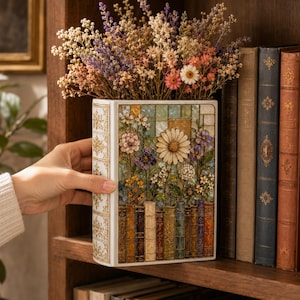 May include: A rectangular ceramic vase shaped like a book with a white spine featuring gold filigree. The front panel displays a mosaic-style illustration of various colourful flowers and a row of vintage book spines at the base, filled with dried flowers.