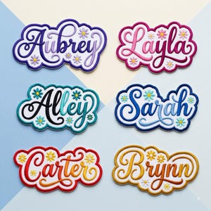 May include: Six embroidered name patches with scalloped edges, each featuring a unique name in cursive script surrounded by small daisy flowers. The names Aubrey, Layla, Alley, Sarah, Carter, and Brynn are displayed in purple, pink, teal, blue, red, and gold.