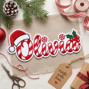 May include: A custom embroidered patch featuring the name Olivia in a red and white candy cane striped font. The letter O is topped with a red Santa hat, and the letter a is accented with a small peppermint candy and green holly leaves.
