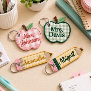 May include: Two apple-shaped keychains with pink and green gingham patterns, embroidered with 'Mrs. Johnson' and 'Mrs. Davis'. Two pencil-shaped keychains with yellow gingham patterns, embroidered with 'Samantha' and 'Abigail'. All have metal rings.