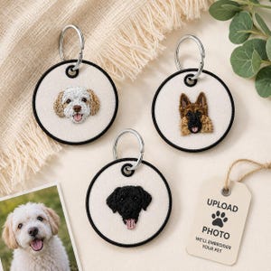 May include: Three circular keychains with off-white fabric centres and black stitched borders. Each features a detailed embroidered dog portrait: a white and tan dog, a German Shepherd, and a black dog. A tag reads UPLOAD PHOTO WE'LL EMBROIDER YOUR PET.