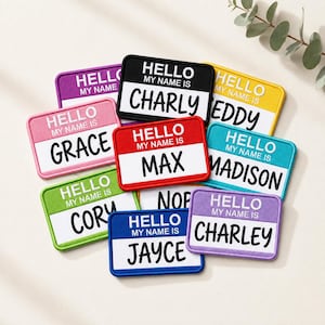 May include: A collection of rectangular name tag patches in various colours including black, red, blue, green, pink, purple, and yellow. Each patch features the text HELLO MY NAME IS in white and black, with a unique name printed below in black font.