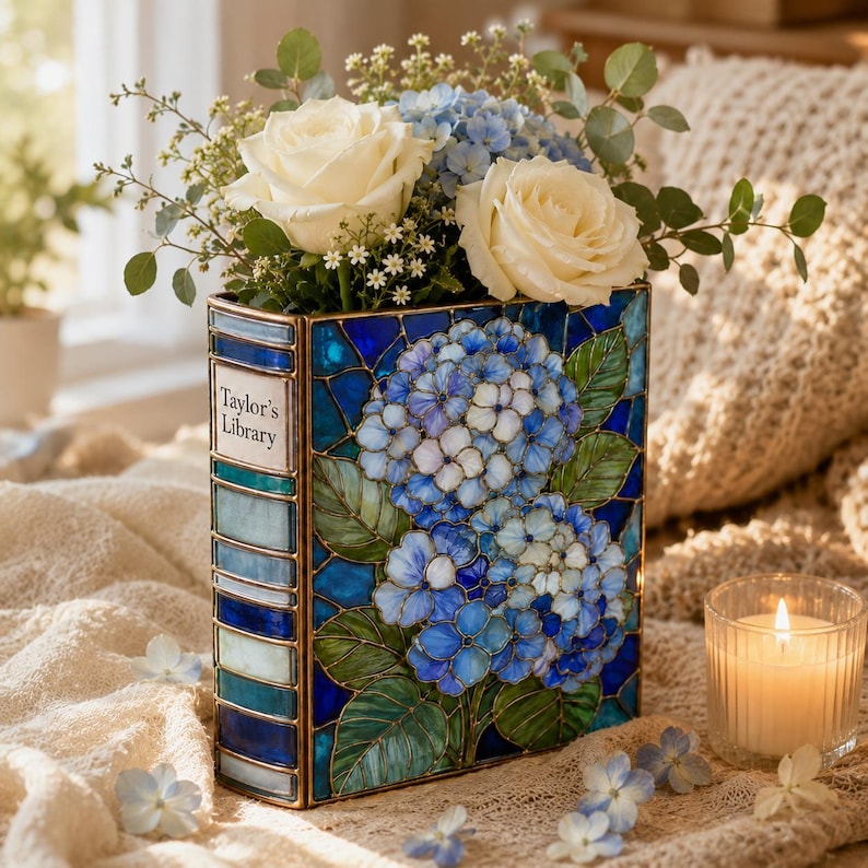 May include: A rectangular stained glass vase shaped like a book with a blue and green hydrangea floral pattern. The spine features the text Taylor's Library. The vase is filled with white roses, blue hydrangeas, and green eucalyptus foliage.