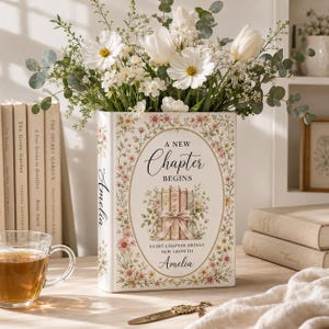 May include: A rectangular white vase shaped like a book, decorated with floral patterns and the text A NEW CHAPTER BEGINS, EVERY CHAPTER BRINGS NEW GROWTH, and the name Amelia. The vase is filled with white cosmos flowers and eucalyptus.