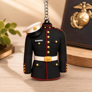 May include: A black military-style dress uniform keychain featuring red trim, gold buttons, and a white belt. A white peaked cap rests on the right shoulder. The name GABRIEL is printed in white on a black rectangular patch above the chest pocket.