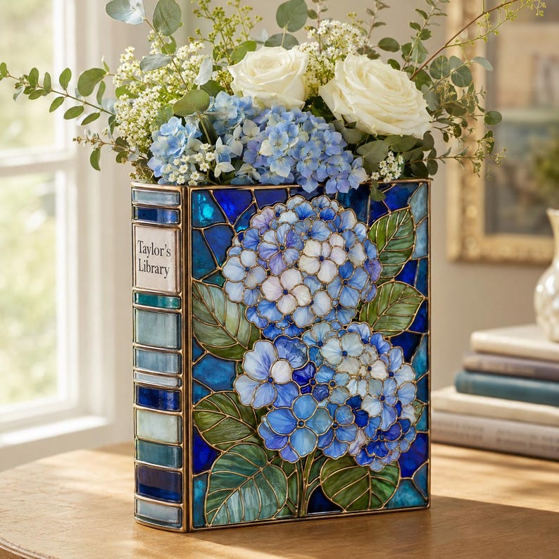 May include: A rectangular stained glass vase shaped like a book with a blue and green hydrangea floral pattern. The spine features the text "Taylor's Library" in white lettering. The vase is filled with white roses and blue hydrangea flowers.