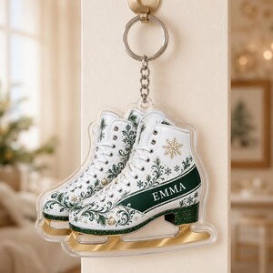 May include: A clear acrylic keychain shaped like an ice skate, featuring a white boot with green and gold floral patterns, a gold blade, and the name EMMA printed in white on a dark green band. It includes a silver metal ring and chain.