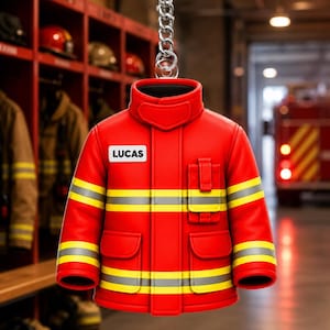 May include: A bright red firefighter jacket keychain featuring horizontal yellow and silver reflective stripes, a small chest pocket, and a white rectangular name tag with the black printed text LUCAS, attached to a silver metal chain.