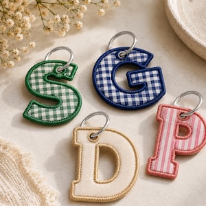 May include: Four fabric letter keychains with metal rings. The letter S features a green and white gingham pattern, the letter G has a blue and white gingham pattern, the letter D is solid beige, and the letter P has a pink and white striped pattern.