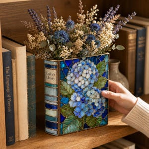 May include: A rectangular vase shaped like a book with the text Taylor's Library on the spine. The vase features a stained glass design of blue and white hydrangea flowers with green leaves, filled with a bouquet of dried blue and beige flowers.