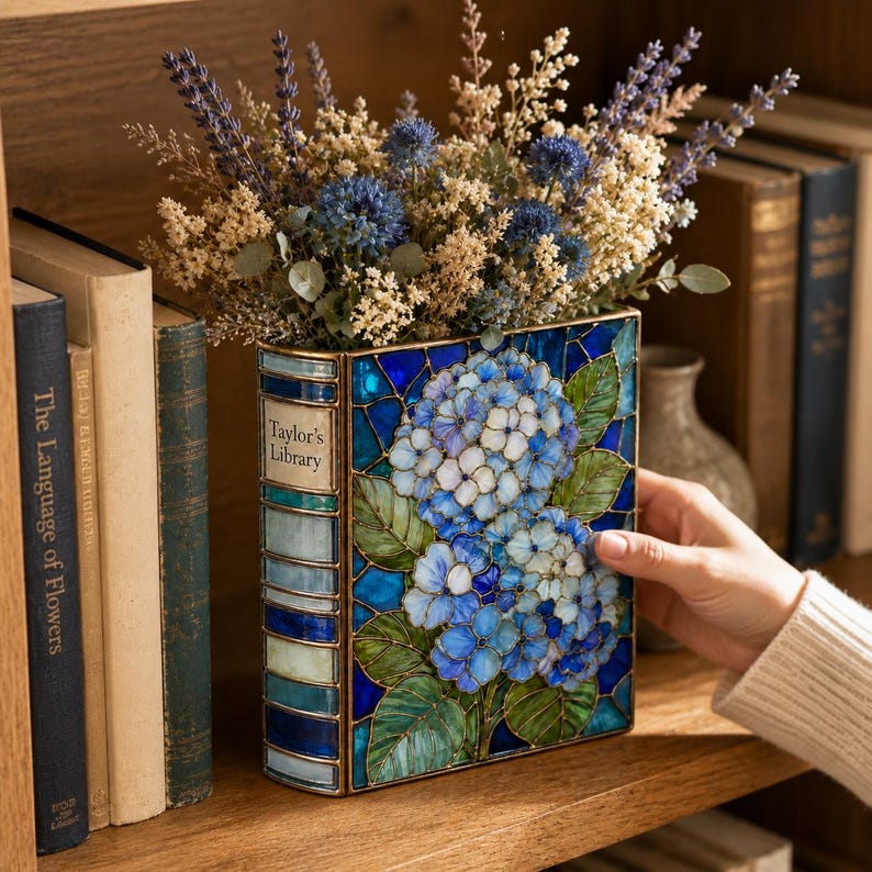 May include: A rectangular vase shaped like a book with the text Taylor's Library on the spine. The vase features a stained glass design of blue and white hydrangea flowers with green leaves, filled with a bouquet of dried blue and beige flowers.