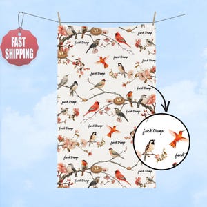 May include: White decorative towel with a repeating pattern of birds, nests, and pink flowers on branches. The words "fuck trump" are written throughout the design. The towel is hanging from a clothesline.