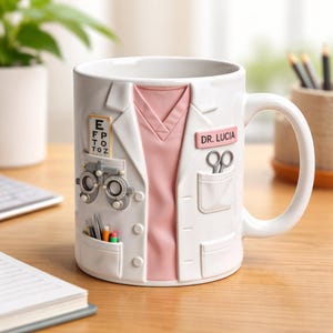 Könnte beinhalten: Weißer Keramikbecher im Design eines Arztkittels, mit einem rosa Hemd und Taschen. Der Becher enthält ein Namensschild mit der Aufschrift "DR. LUCIA", eine Sehtafel und eine Schere. Ein einzigartiges Geschenk für medizinische Fachkräfte.