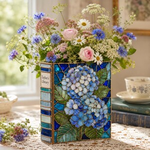 May include: A rectangular vase shaped like a book with a stained glass design featuring blue hydrangea flowers and green leaves. The spine is labeled Taylor's Library. The vase is filled with a bouquet of blue cornflowers, pink roses, and white daisies.