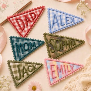 May include: Six triangular fabric pennants with scalloped edges, each featuring embroidered names or titles. The designs include red gingham with DAD, blue gingham with ALEX, teal gingham with MOM, camouflage with SOPHIA, olive gingham with JACK, and pastel plaid with EMILY.