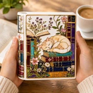 May include: A white ceramic book-shaped box featuring a colorful stained-glass style illustration of a sleeping cat resting on a stack of books, surrounded by floral motifs, with gold-toned outlines and a vertical spine design on the left side.