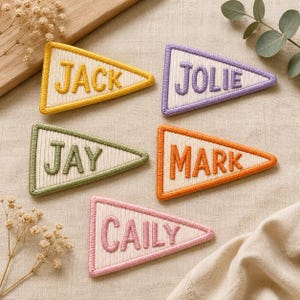 May include: Five triangular embroidered patches with a ribbed cream-colored center and colored borders. The patches feature the names JACK in yellow, JOLIE in purple, JAY in green, MARK in orange, and CAILY in pink, all stitched in capital letters.