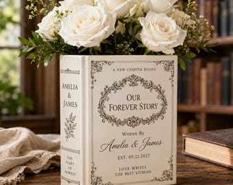 May include: A cream-coloured ceramic book-shaped vase filled with white roses and greenery. The front features black text reading A NEW CHAPTER BEGINS, OUR FOREVER STORY, Written By Amelia & James, EST. 09.21.2027, LOVE WRITES THE BEST STORIES.