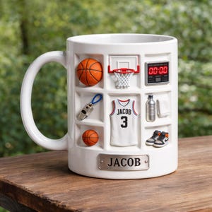 Può includere: Tazza in ceramica bianca con decorazioni a tema basket. Presenta un pallone da basket, un canestro, una maglia con il nome "JACOB" e il numero 3, un timer, scarpe da ginnastica e una bottiglia d'acqua. Include una targhetta metallica con il nome "JACOB".