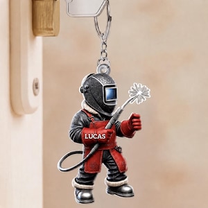 May include: A keyring featuring a cartoon welder wearing a black and red protective suit with a welding helmet. The figure holds a welding torch emitting a starburst spark. The name LUCAS is printed in white on the red chest apron of the suit.