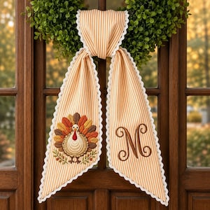 May include: A beige and white striped fabric door hanger featuring a white scalloped lace trim. One side displays an embroidered turkey with colorful feathers, while the other side features a large, dark brown embroidered letter M monogram.