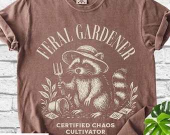 Può includere: T-shirt color espresso con un procione con un cappello che tiene un forcone da giardinaggio. Il testo "FERAL GARDENER" è sopra il procione, con "CERTIFIED CHAOS CULTIVATOR" sotto. Il design include bustine di semi e un piccolo vaso.