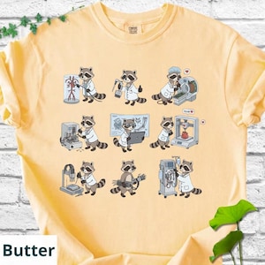 May include: Butter-colored t-shirt featuring a repeating pattern of raccoons in lab coats, each engaged in different scientific activities. The raccoons are depicted with various medical equipment and tools, creating a playful and educational design.