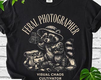 May include: Black t-shirt with a raccoon graphic wearing a hat and glasses, holding cameras. The text reads "FERAL PHOTOGRAPHER" above the raccoon and "VISUAL CHAOS CULTIVATOR" below.
