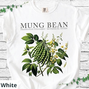 May include: White t-shirt featuring a botanical illustration of mung beans. The design includes green mung bean pods, leaves, and yellow flowers, with the text "MUNG BEAN" and "PHASEOLUS VULGARIS - BOTANICAL STUDY".