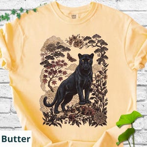 May include: Butter-colored t-shirt featuring a black panther illustration. The graphic includes stylized trees, flowers, and clouds, with a butterfly accent. The shirt has a classic crew neck and short sleeves.