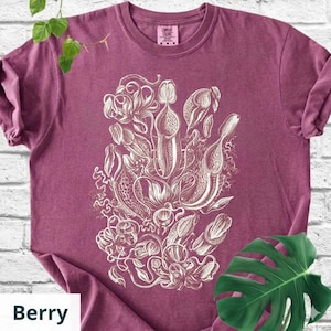 May include: Berry-colored t-shirt featuring a detailed white botanical illustration of various pitcher plants. The design includes intricate line work and shading, creating a textured appearance. The shirt has a classic crew neck and short sleeves.