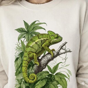 May include: Cream-colored sweatshirt featuring a detailed illustration of a green chameleon perched on a branch, surrounded by lush green leaves. The design has a vintage botanical art style.