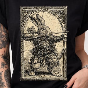 May include: Black t-shirt featuring a detailed illustration of a rabbit archer aiming a bow and arrow. The design is in a vintage style with intricate linework and a medieval aesthetic. The rabbit is dressed in armor.