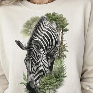 May include: Cream-colored sweatshirt featuring a detailed black and white illustration of a zebra grazing. The zebra is set against a backdrop of green foliage and trees, creating a nature-inspired design.