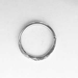 May include: A silver-colored, hammered metal ring with a hinged clasp. The ring is circular with a textured surface, and the clasp is located on the outer edge. The ring is set against a white background.