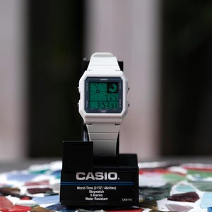 Puede incluir: Un reloj digital Casio blanco con una pantalla verde que muestra la hora y la fecha. El reloj está sobre un soporte negro con el logotipo de Casio y funciones como hora mundial, cronómetro y alarmas. El reloj es resistente al agua.