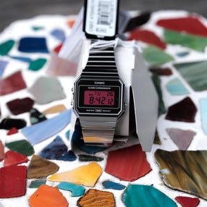 May include: A Casio digital watch with a black face and silver band. The watch displays the time, date, and alarm settings in red. The watch is new, with the original tag attached. The watch is displayed on a white surface with colorful mosaic tiles.