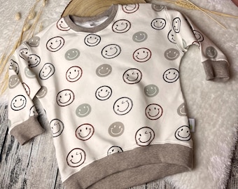May include: Cream-colored sweatshirt featuring a repeating pattern of smiley faces in black, brown, and gray. The sweatshirt has brown cuffs, neckline, and hem. The garment is displayed on a white fluffy surface.
