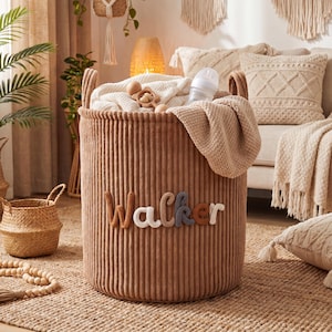 Op de afbeelding: Een cilindervormige opbergmand van bruin corduroy met de naam Walker in verhoogde, meerkleurige gebreide letters op de voorzijde. De mand is gevuld met een witte gebreide deken, een houten rammelaar en een doorzichtige plastic babyfles.