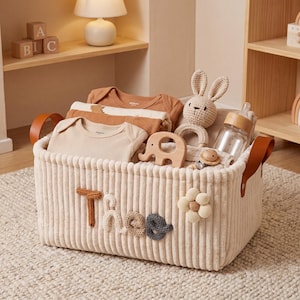 Könnte beinhalten: Ein cremefarbener Cord-Aufbewahrungskorb mit dem Namen "Theo" in strukturierten Buchstaben. Der Korb enthält Babykleidung, ein Stoffkaninchen, ein Holz-Elefantenspielzeug, eine Babyflasche und eine Blumenapplikation.