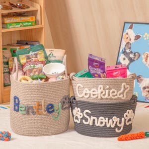 May include: Three woven rope pet baskets: a tan one with "Bentley" in colourful letters, a tan one with "Cookie" and a bone, and a grey one with "Snowy" and paw prints. The baskets are filled with pet supplies.