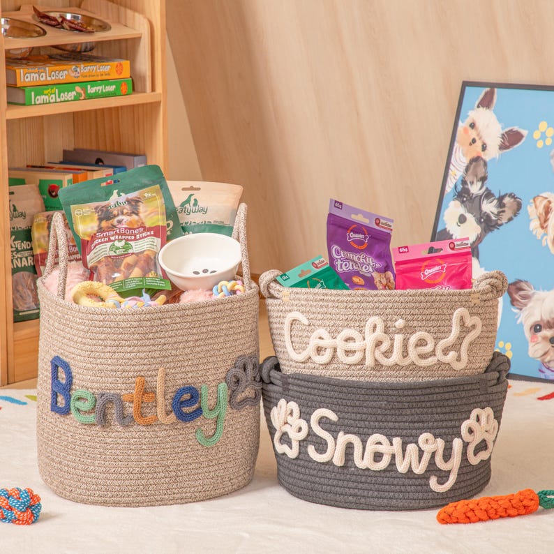 May include: Three woven rope pet baskets: a tan one with "Bentley" in colourful letters, a tan one with "Cookie" and a bone, and a grey one with "Snowy" and paw prints. The baskets are filled with pet supplies.