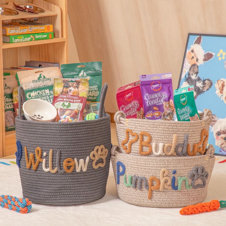 May include: Three woven rope baskets for pets, filled with treats and toys. The gray basket is labelled "Willow" with a paw print, the beige basket says "Buddah", and the bottom basket says "Pumpkin" with a paw print. A white bowl is inside the gray basket.