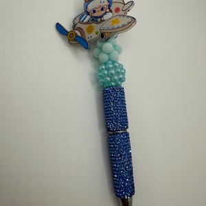 May include: A blue pen with a decorative top featuring a cartoon pilot in a biplane. The pen's barrel is covered in sparkling blue rhinestones, and the top has light blue pom-poms and beads.