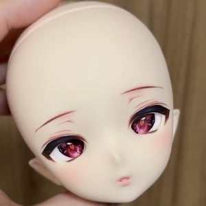 Pode incluir: Cabeça de boneca com tez pálida, com olhos grandes e expressivos com íris rosa escuro e delineador preto. A boneca tem um rubor sutil nas bochechas e sobrancelhas delicadas pintadas. Os lábios são rosa suave.
