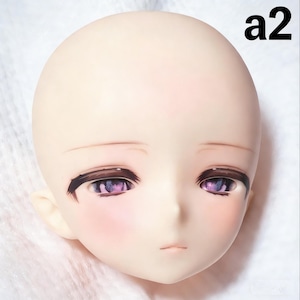 May include: A doll head with a smooth, pale complexion. The eyes are detailed with purple irises and dark brown eyeliner. The cheeks have a subtle pink blush, and the lips are a pale pink. The text "a2" is in the upper right corner.