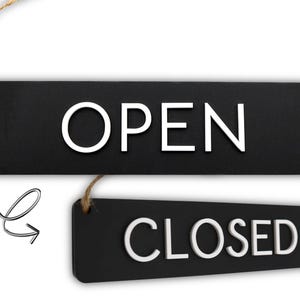 Op de afbeelding: Twee zwarte rechthoekige borden met witte tekst en touwhangers. Het bovenste bord leest "OPEN", en het onderste bord leest "CLOSED". Een decoratieve krul en pijl staan in de linker benedenhoek. De borden zijn waarschijnlijk voor een bedrijf.