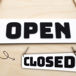 May include: Two rectangular signs, one reading "OPEN" and the other "CLOSED", in bold black letters on a white background. Both signs are suspended by natural rope, suitable for business use.